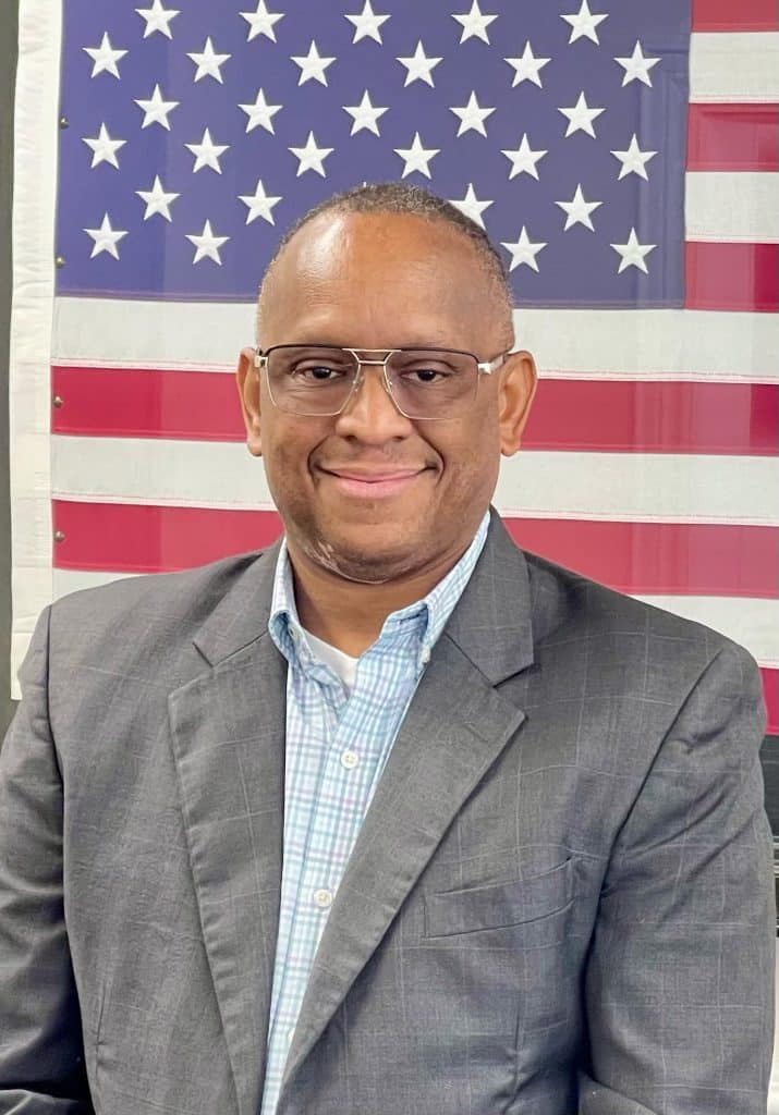 Smiling man in suit with American flag backdrop.